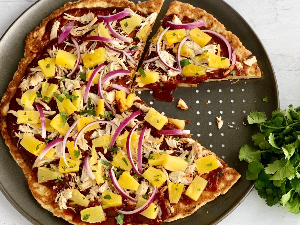 A Hawaiian pizza with BBQ sauce, shredded chicken, purple onions pieces, and diced pineapple.