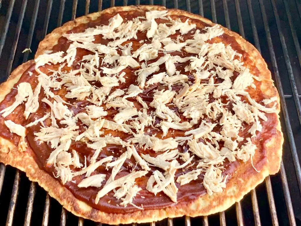 A Hawaiian pizza with BBQ sauce, shredded chicken being cooked.