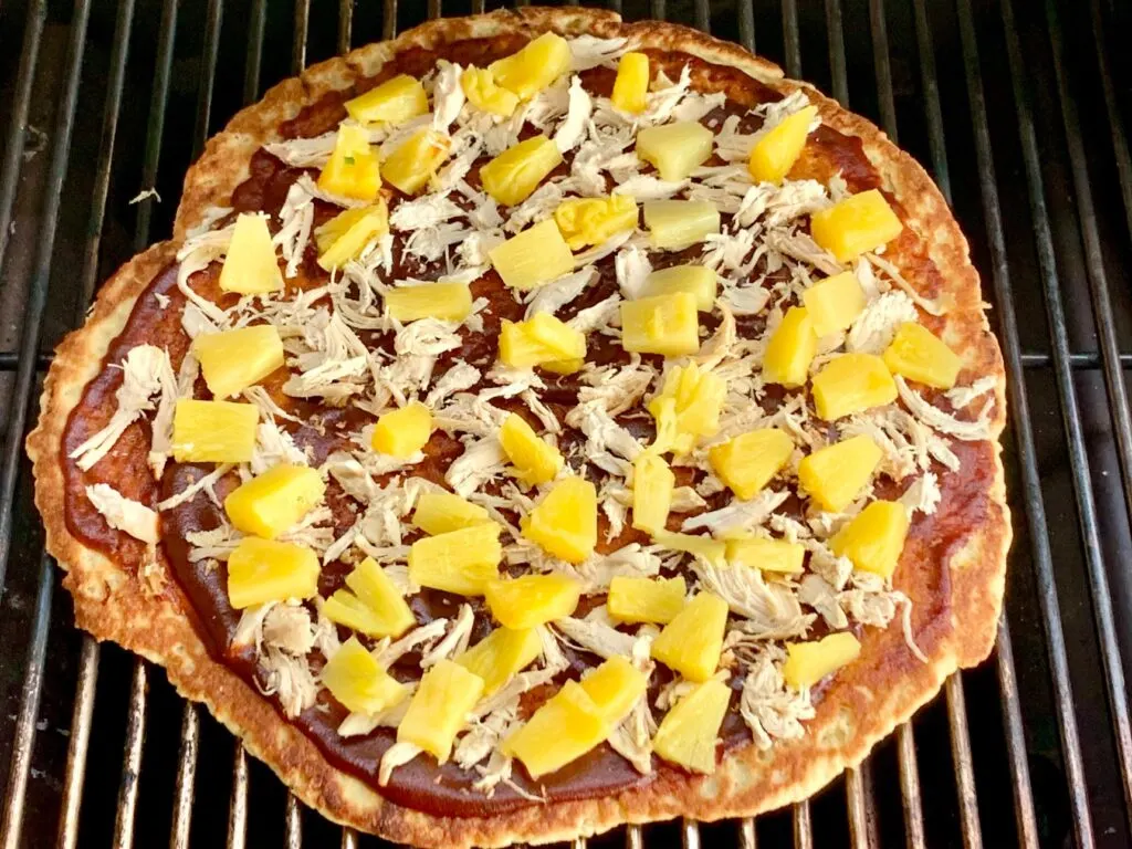 A Hawaiian pizza with BBQ sauce, shredded chicken, and diced pineapple on a grill being cooked.