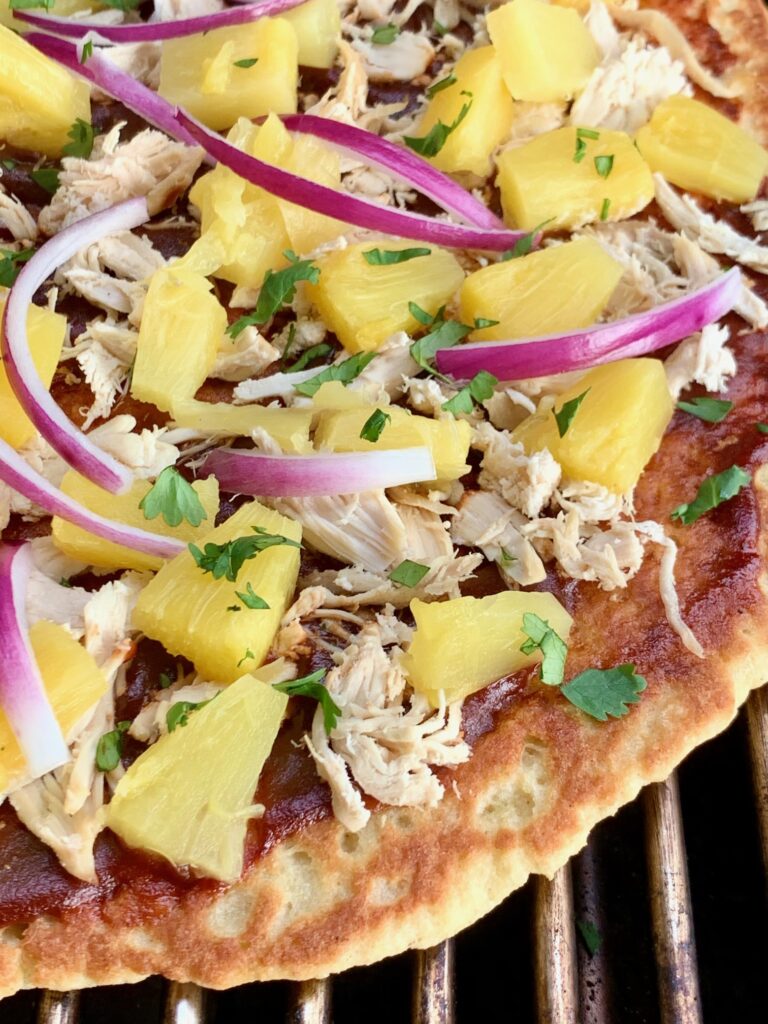 A Hawaiian pizza with BBQ sauce, shredded chicken, purple onions pieces, and diced pineapple on a grill being cooked.