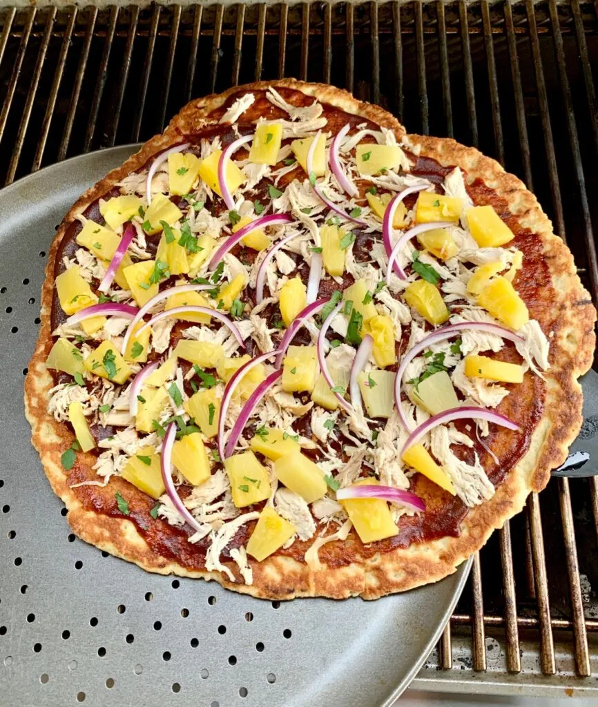 A Hawaiian pizza with BBQ sauce, shredded chicken, purple onions pieces, and diced pineapple on a grill being transferred to a pizza pan.