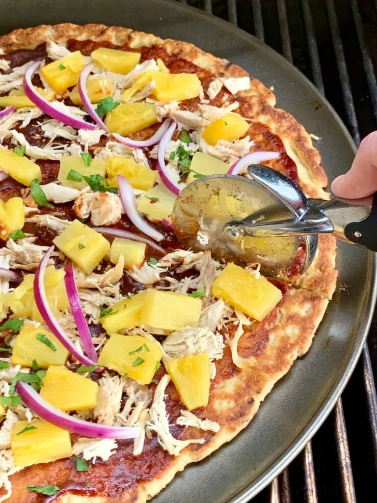 A Hawaiian pizza with BBQ sauce, shredded chicken, purple onions pieces, and diced pineapple on a grill being cut with a pizza cutter.