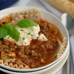 Several breadsticks next to a large soup bowl of creamy lasagna soup. This soup features mushrooms, tender gluten free lasagna noodles, and dairy free ricotta.