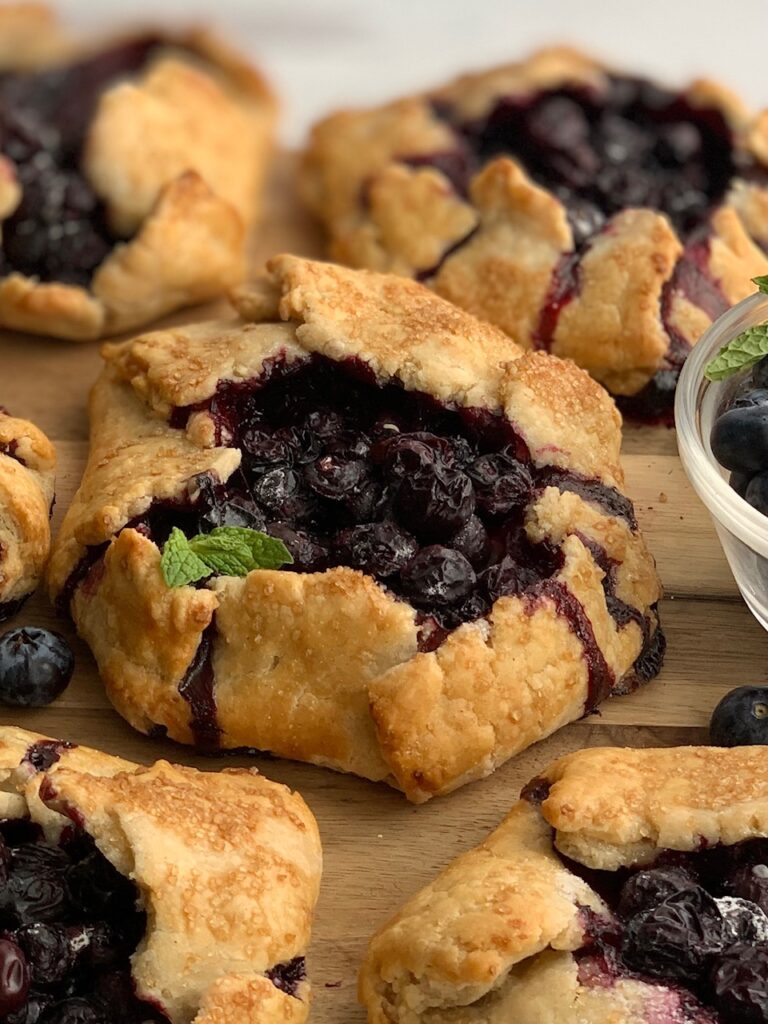 A rustic version of a blueberry pie. A flat pie crust with formed sides to hold a blueberry filling surrounded by more galettes on all sides. On top is a fresh mint leave.