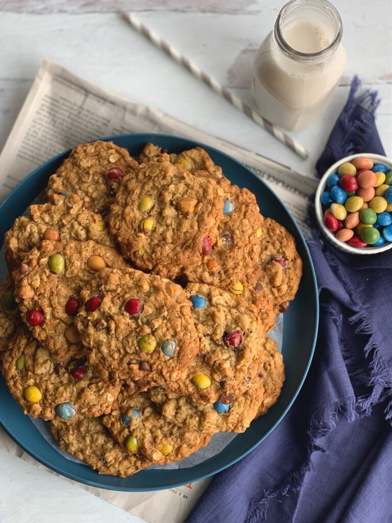 An overhead shot of a blue plate full of cookies with oats, creamy peanut butter, chocolate morsels, and chocolate candies next to a glass of milk.