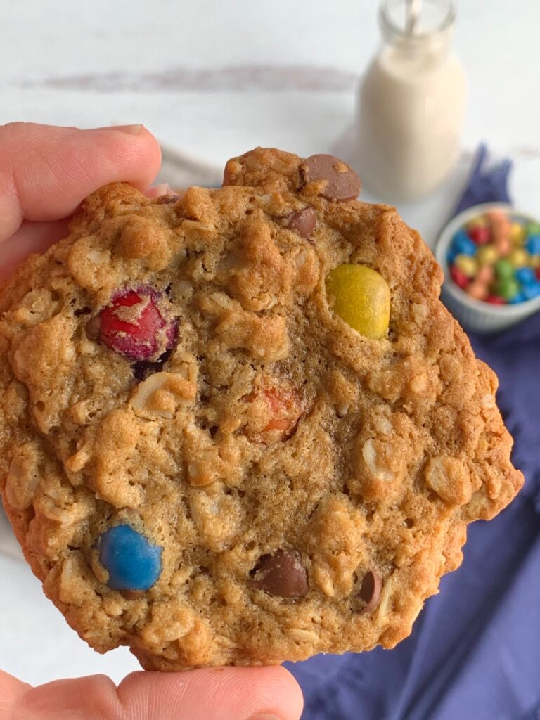 A hand holding a cookie with oats, creamy peanut butter, chocolate morsels, and chocolate candies.