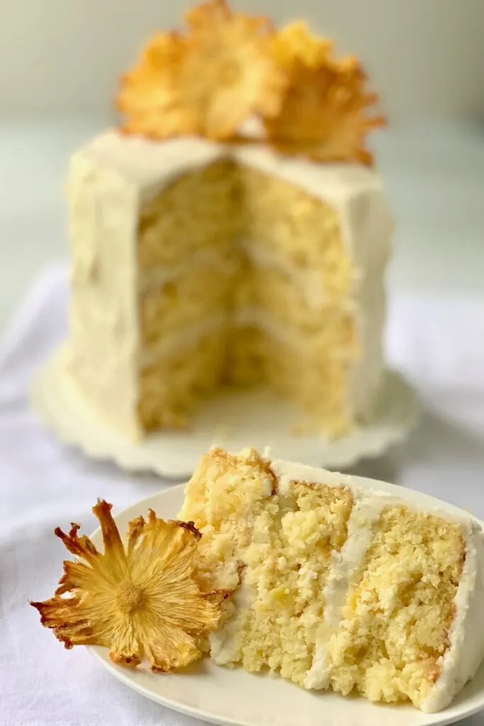 A large slice of cake with small pineapple chunks and  buttercream frosting and a small 3 tired cake behind it with dried pineapple rings on it.