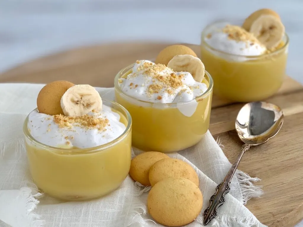 3 mini dessert glasses filled with layers of sliced bananas, vanilla wafer cookies, banana pudding, and creamy whipped topping. Then cookie crumbs, another cookie, and a banana slice on top of that.