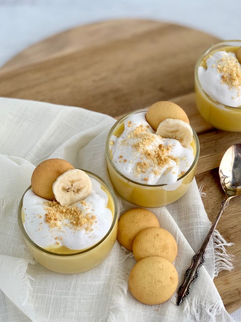 The top view looking down of 3 mini dessert glasses filled with layers of sliced bananas, vanilla wafer cookies, banana pudding, and creamy whipped topping. Then cookie crumbs, another cookie, and a banana slice on top of that.