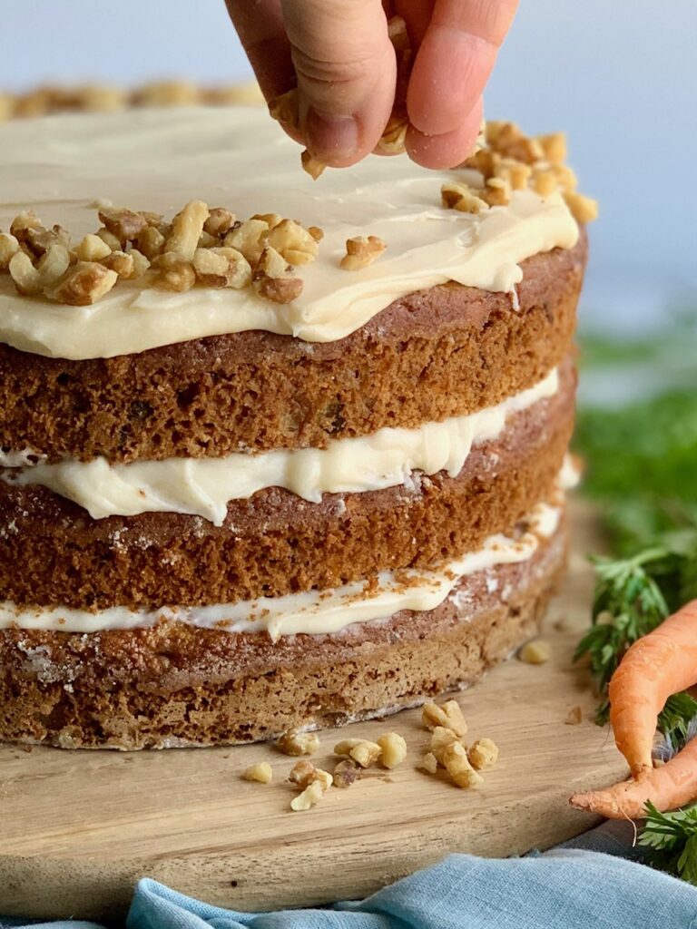 A 3 tiered carrot cake with a white cream cheese frosting in-between each layer and on top with a hand shown sprinkling nuts over the top.