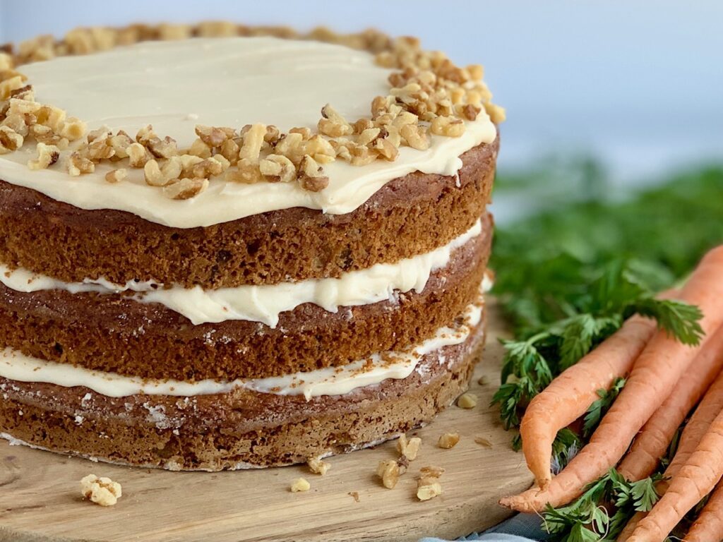 A 3 tiered carrot cake with a white cream cheese frosting in-between each layer and on top with nuts next to a bunch of fresh carrots with long green tops.