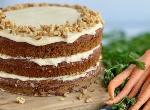 A 3 tiered carrot cake with a white cream cheese frosting in-between each layer and on top with nuts. A slice has been cut out of the cake and is being lifted up by a serving utensil.