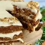 A 3 tiered carrot cake with a white cream cheese frosting in-between each layer and on top with nuts. A slice has been cut out of the cake and is being lifted up by a serving utensil.