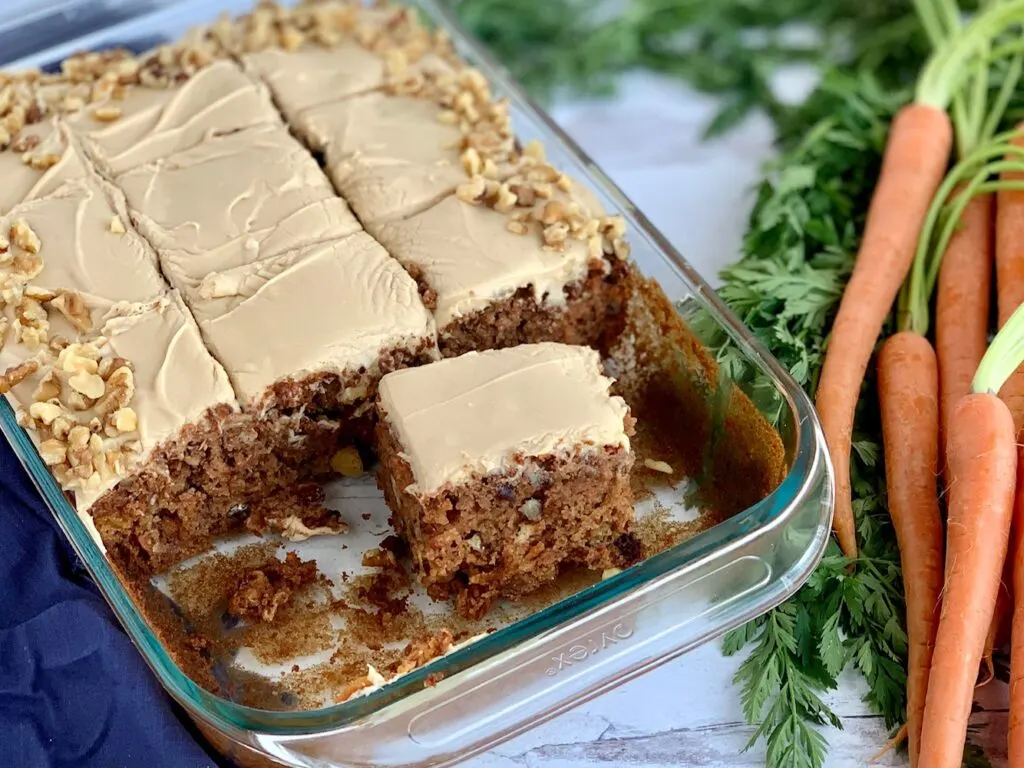 A 9 x 13" cake pan filled with a shredded cake mixture with unsweetened applesauce, crushed pineapple, and topped with a creamy brown sugar cream cheese frosting and chopped walnuts around the outside edge of the cake next to a bunch of carrots. A few pieces of cake have been removed to show the inside of the cake.