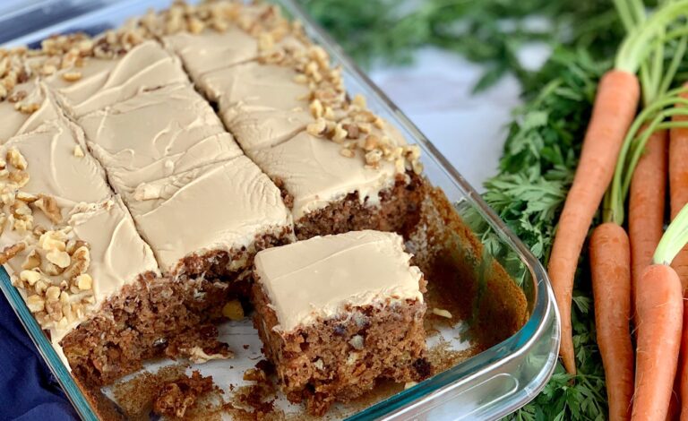 A 9 x 13" cake pan filled with a shredded cake mixture with unsweetened applesauce, crushed pineapple, and topped with a creamy brown sugar cream cheese frosting and chopped walnuts around the outside edge of the cake next to a bunch of carrots. A few pieces of cake have been removed to show the inside of the cake.