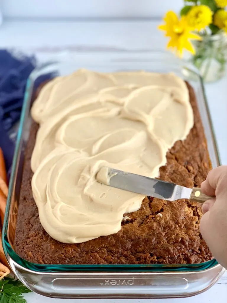 A moist cake being frosted with a small angled spatula pushing a brown sugar cream cheese frosting around cake. 