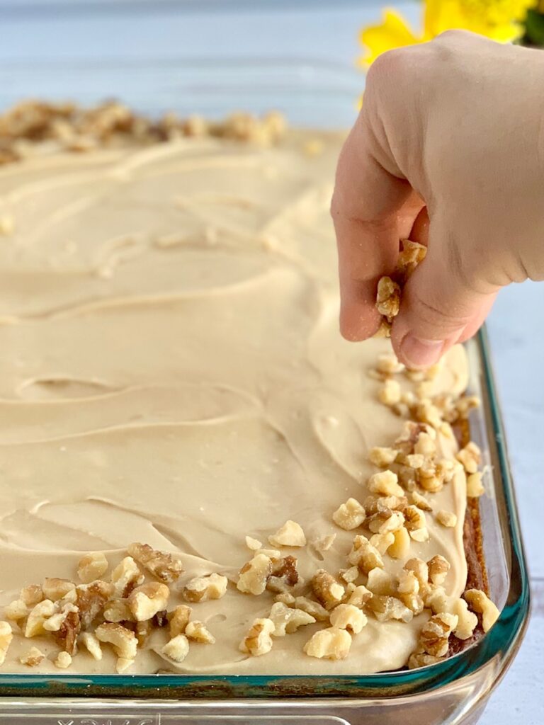 A cake that's now frosted and you see a hand dropping chopped walnuts around the outside edge of it. 