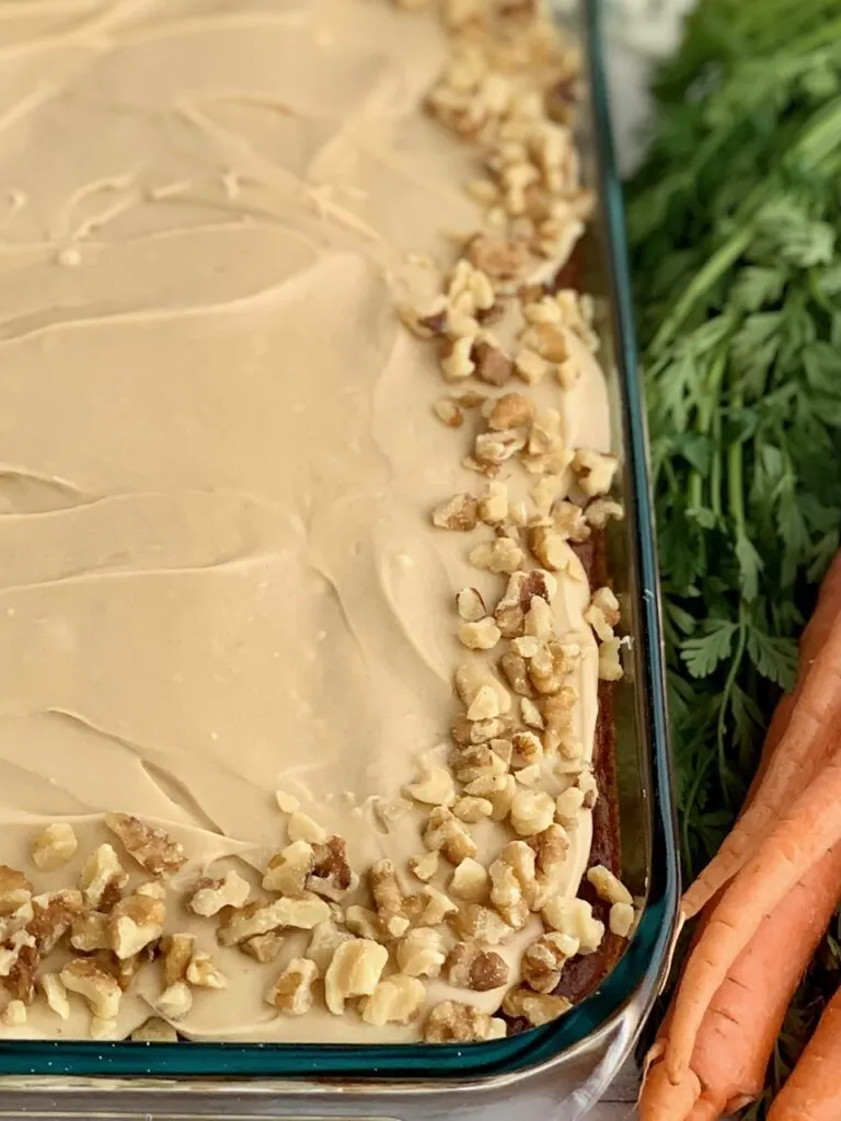 A 9 x 13" baking pan filled with a shredded cake mixture with unsweetened applesauce, crushed pineapple, and topped with a creamy brown sugar cream cheese frosting and chopped walnuts around the outside edge of the cake next to a bunch of carrots.