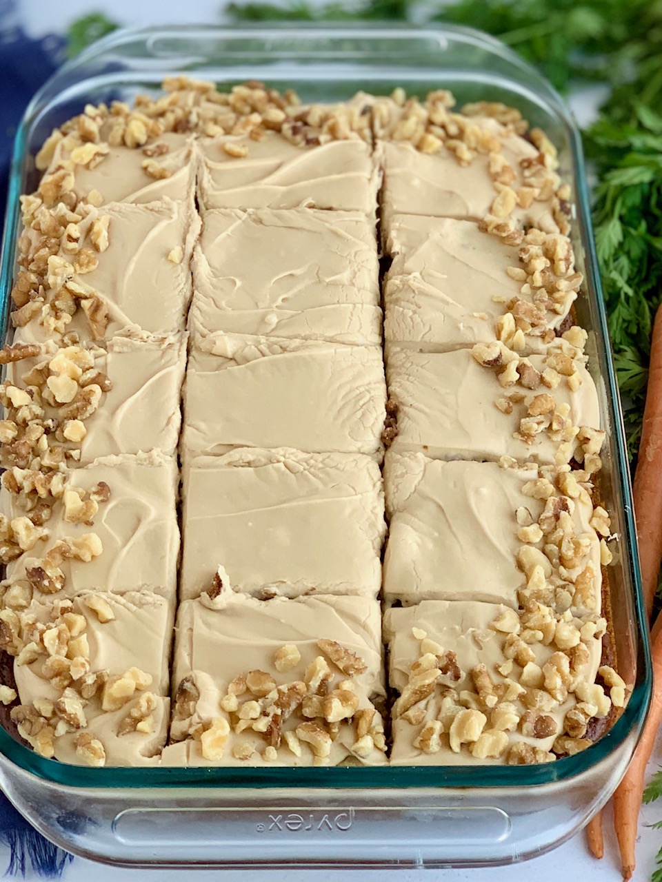 A 9 x 13" baking pan filled with a shredded cake mixture with unsweetened applesauce, crushed pineapple, and topped with a creamy brown sugar cream cheese frosting and chopped walnuts around the outside edge of the cake next to a bunch of carrots.