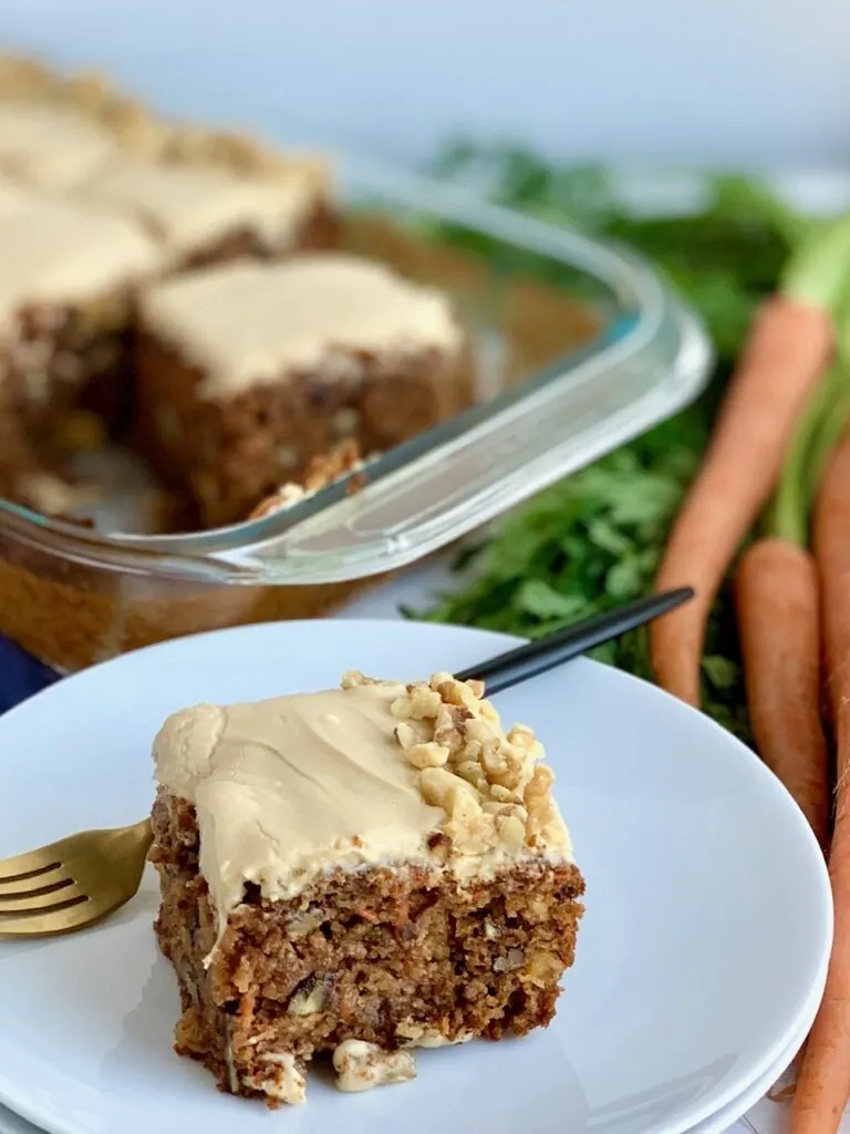 A 9 x 13" baking pan filled with a shredded cake mixture with unsweetened applesauce, crushed pineapple, and topped with a creamy brown sugar cream cheese frosting and chopped walnuts around the outside edge of the cake next to a bunch of carrots. And a slice of cake on a white cake plate next to a fork.