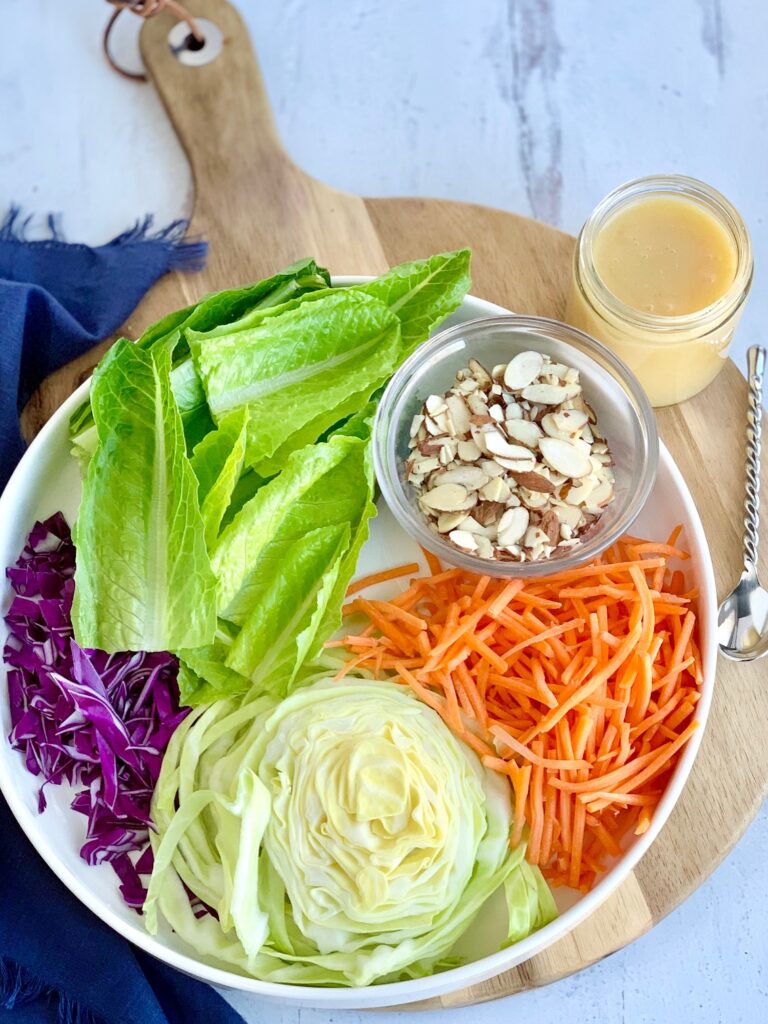 A platter with a small bowl of sliced almonds, a grouping of shredded carrots next to a cutting of light green napa cabbage, and to the left of that is small cuttings of purple cabbage next to large strips of romaine lettuce. To the right of the serving platter is a small spoon and a glass container of dressing.
