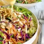 A large salad in a serving platter. The salad has cuts of romaine lettuce, purple cabbage, light green napa cabbage, chunks of grilled chicken, shredded carrots, and lots of sliced almonds. There is even a few strips of wonton strips. Over the top of the salad is a dressing being poured onto it.