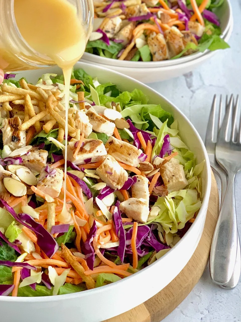 A large salad in a serving platter. The salad has cuts of romaine lettuce, purple cabbage, light green napa cabbage, chunks of grilled chicken, shredded carrots, and lots of sliced almonds. There is even a few strips of wonton strips. Over the top of the salad is a dressing being poured onto it.