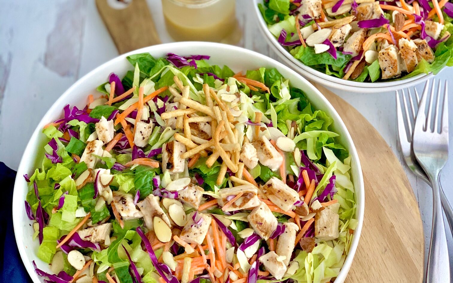 A large salad in a serving platter. The salad has cuts of romaine lettuce, purple cabbage, light green napa cabbage, chunks of grilled chicken, shredded carrots, and lots of sliced almonds. There is even a few strips of wonton strips.