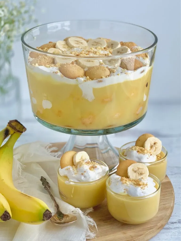 3 mini dessert glasses filled with layers of sliced bananas, vanilla wafer cookies, banana pudding, and creamy whipped topping. Then cookie crumbs, another cookie, and a banana slice on top of that next to a trifle bowl filled with the same layers and toppings.