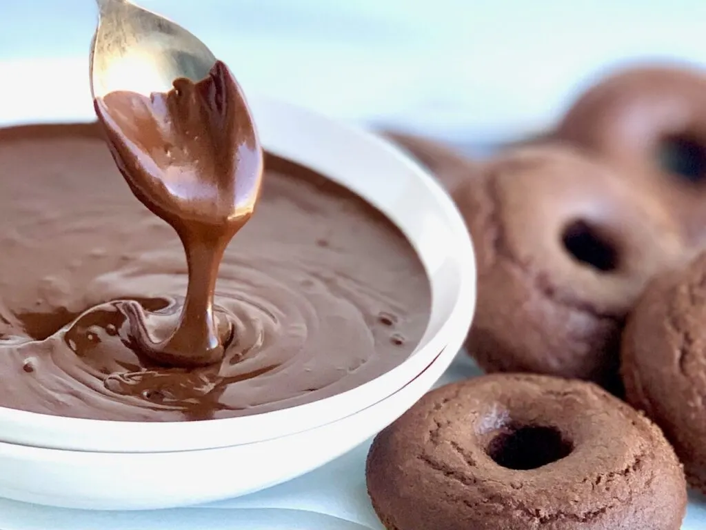 A white bowl holding warm and runny chocolate ganache dripping off of a spoon next to several chocolate donuts. 