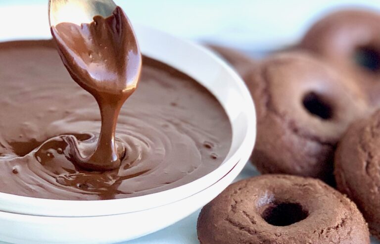 A white bowl holding warm and runny chocolate ganache dripping off of a spoon next to several chocolate donuts.