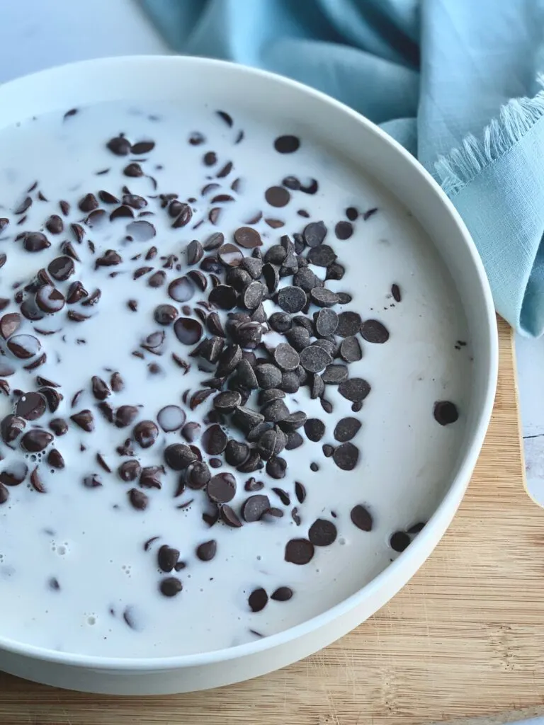 A white serving dish with dairy free 
 chocolate morsels and a heated coconut cream mixture.