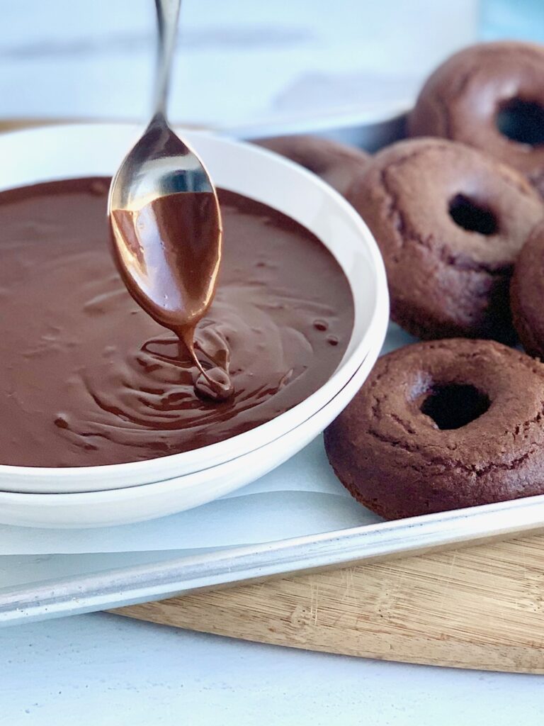 A white bowl holding warm and runny chocolate ganache dripping off of a spoon next to several chocolate donuts. 