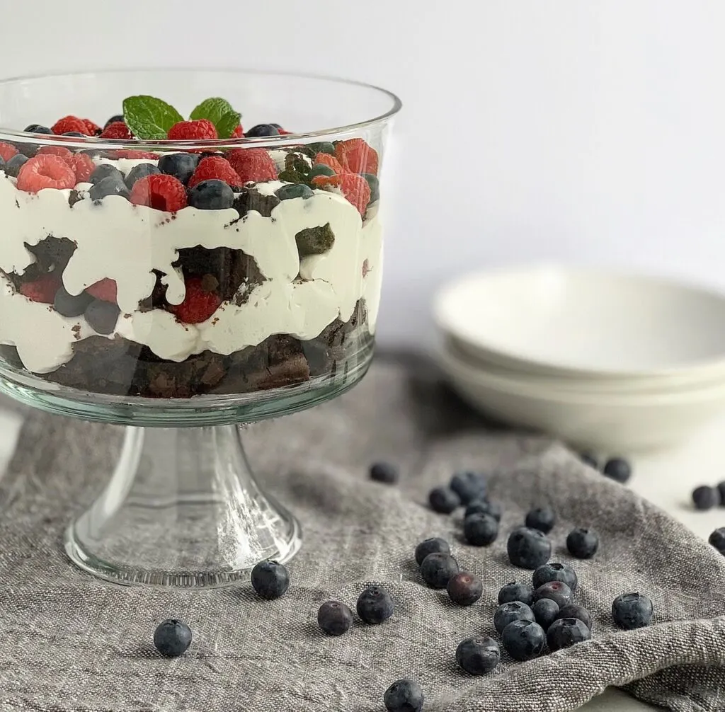 A large glass trifle bowl with brownie chunks, creamy cocowhip, blueberries and raspberries in it,