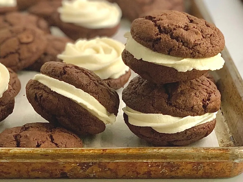 A baking sheet full of small brownie cookies with buttercream icing in-between them to for a brownie cookie sandwich,,