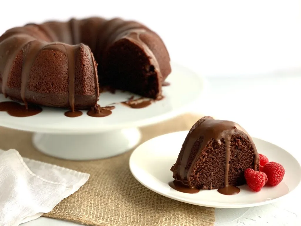 A round chocolate bundt cake with a couple of slices cut out of it. One slices is on a plate next to it with chocolate ganache running down it and fresh raspberries.