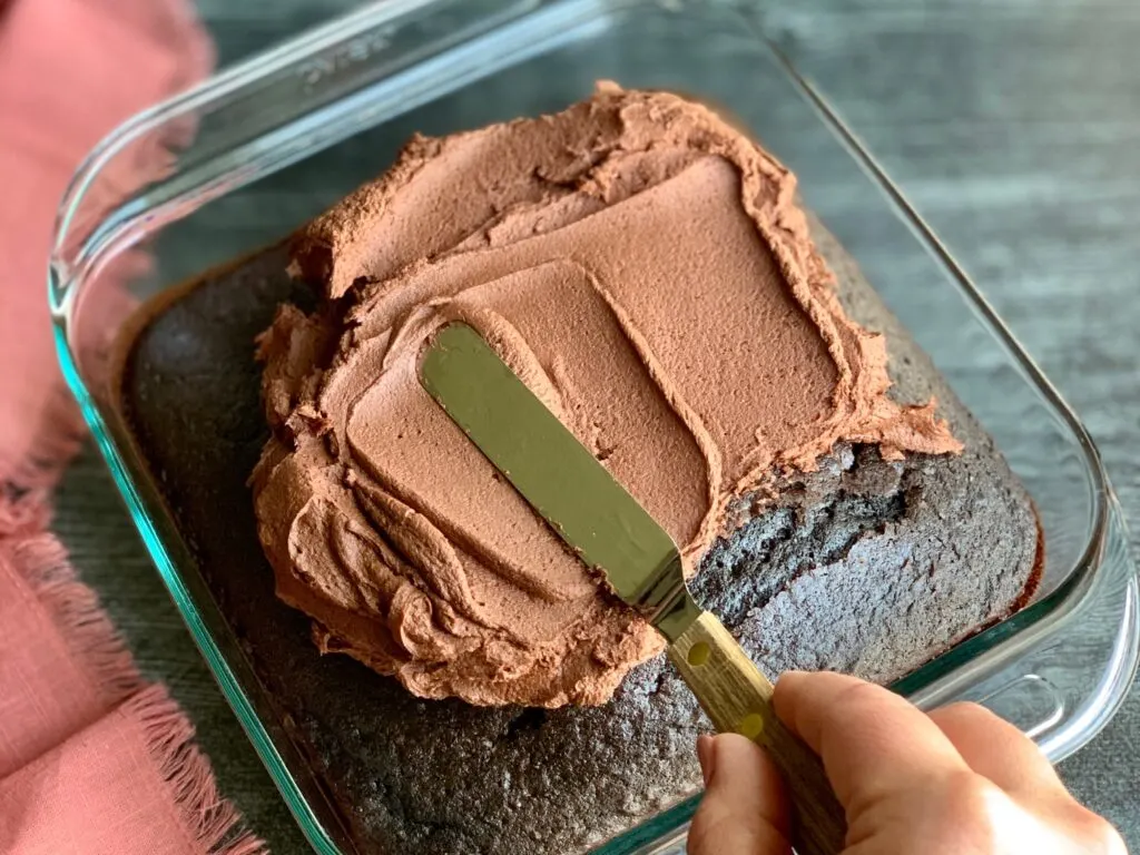 A glass pan holding a chocolate cake. An angled spatula is spreading chocolate buttercream frosting over the top of the cake.