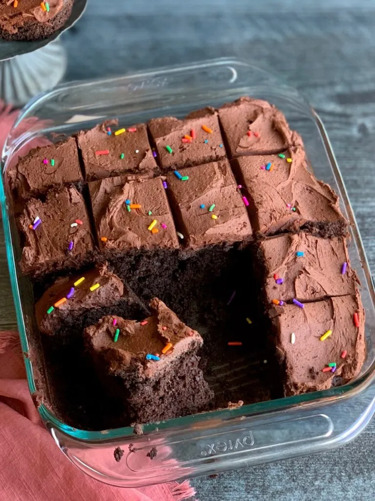 A glass pan holding a chocolate cake frosted with chocolate buttercream and topped with colorful sprinkles. A few pieces of the cut cake have been removed.