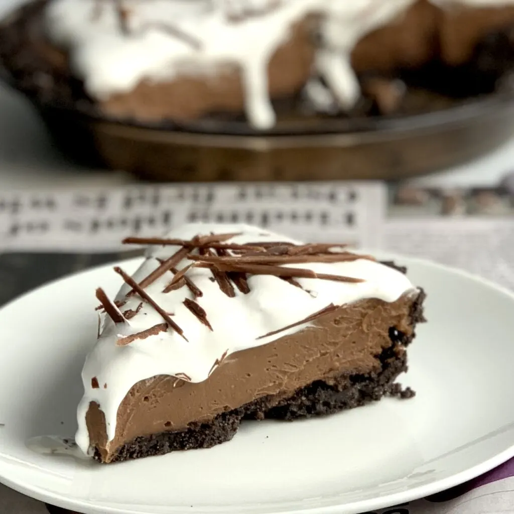 A  slice of chocolate cream pie. The first layer is an oreo like cookie crust followed by a thick chocolate pudding then topped with cocowhip and decorative chocolate shavings. the rest of the pie is in the background,