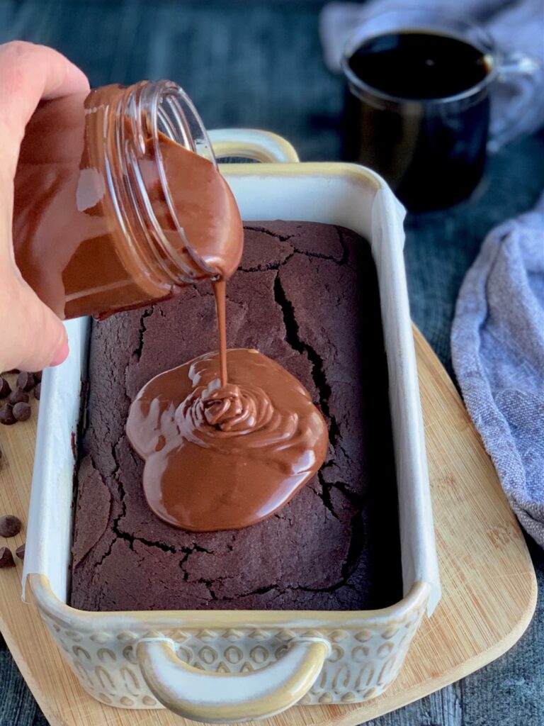 A bread loaf pan with a fresh baked chocolate loaf in it with someone drizzling fresh chocolate ganache on top next to a few scattered chocolate chips.