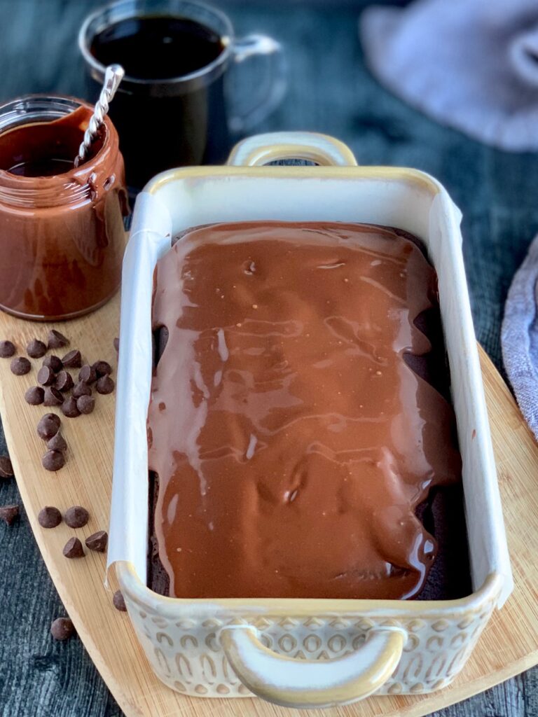 A bread loaf pan with a fresh baked chocolate loaf in it and smooth chocolate ganache frosting next to a few scattered chocolate chips.