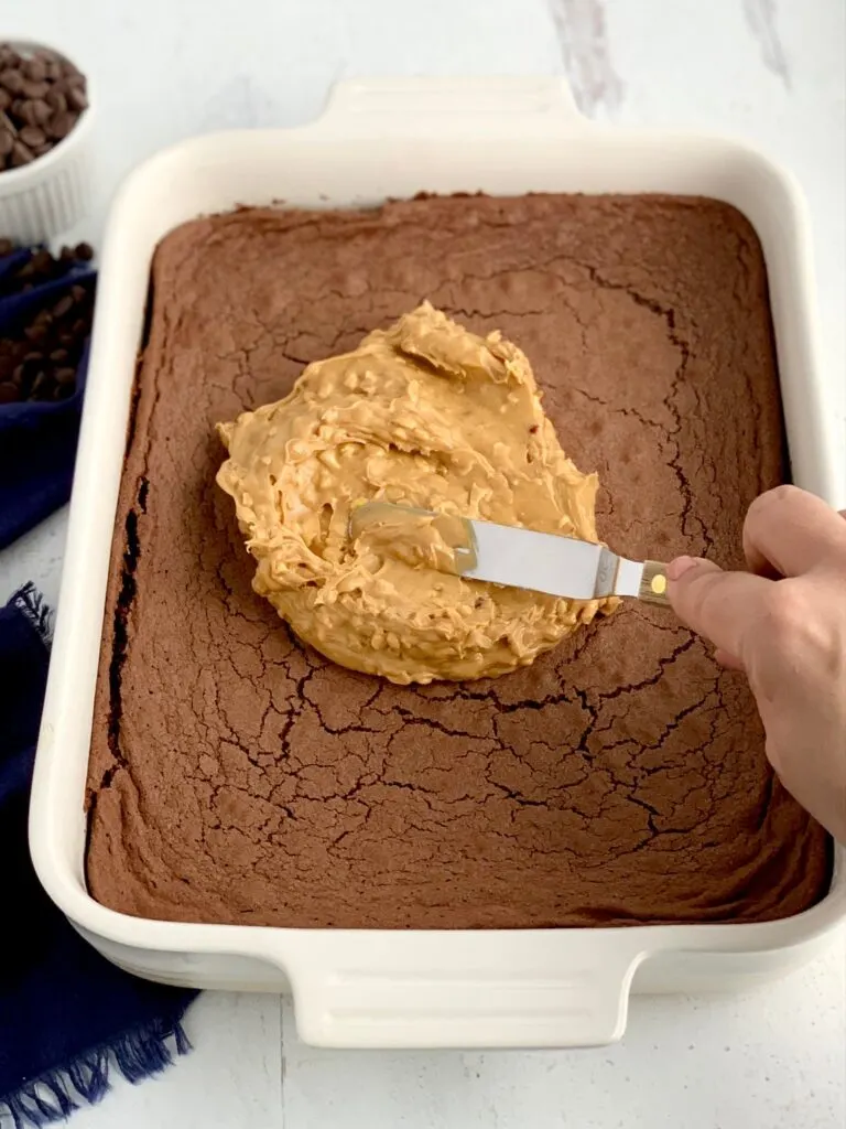 A baked chocolate brownie mix in a white 9X13-inch baking pan and someone spreading crunchy peanut butter over the cooled brownie.