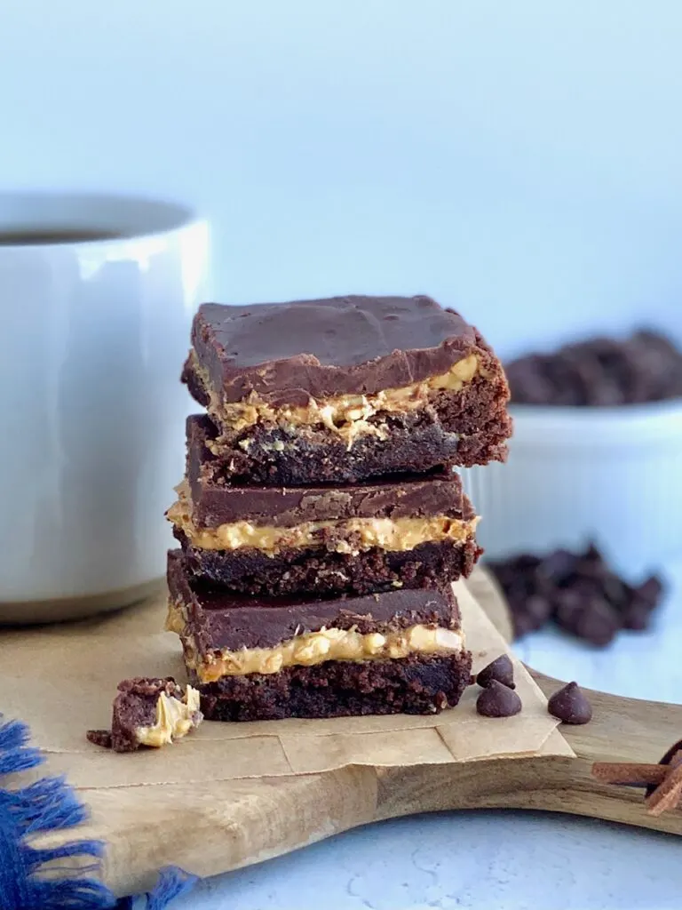 A stack of 3 cut bars. The first layer of the bars is a chocolate brownie, with crunchy peanut butter slathered over it, then topped with a soft chocolate marshmallow mixture next to a cup of coffee.