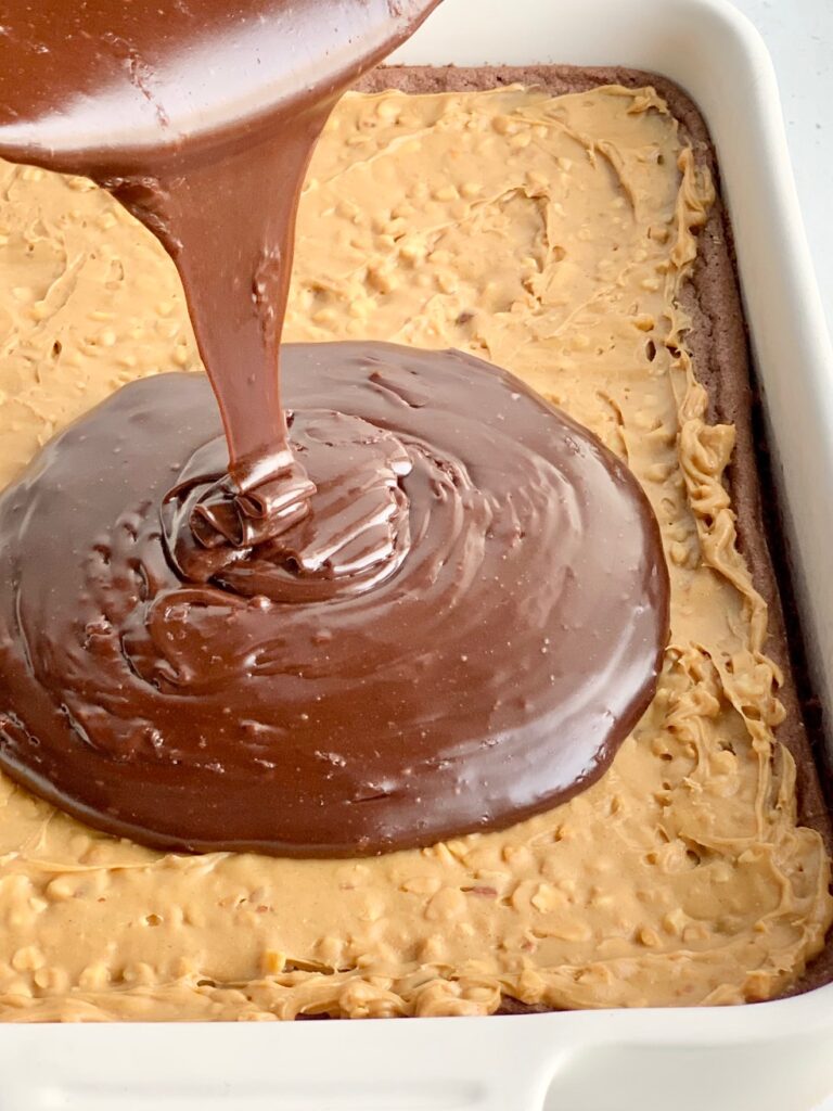 A baked chocolate brownie mix in a white 9X13-inch baking pan with crunchy peanut butter over the cooled brownie. Someone is pouring a warm chocolate mixture over it.