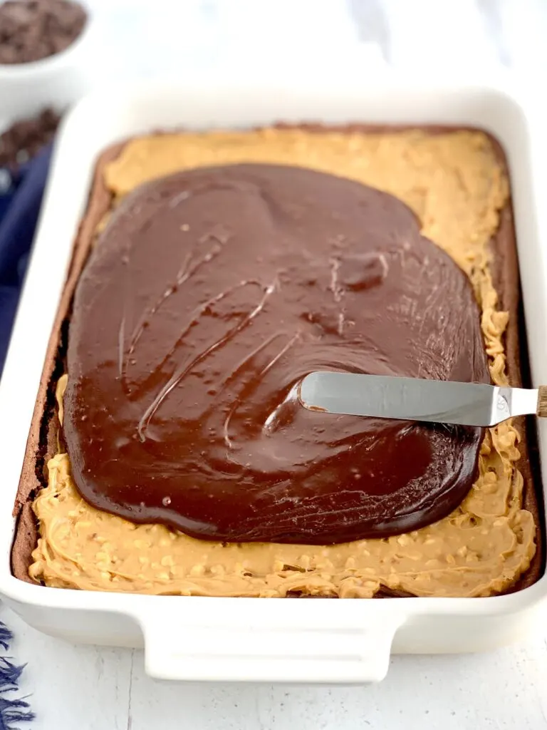 A baked chocolate brownie mix in a white 9X13-inch baking pan with crunchy peanut butter over the cooled brownie. Someone is spreading a warm chocolate mixture over the crunchy peanut butter.