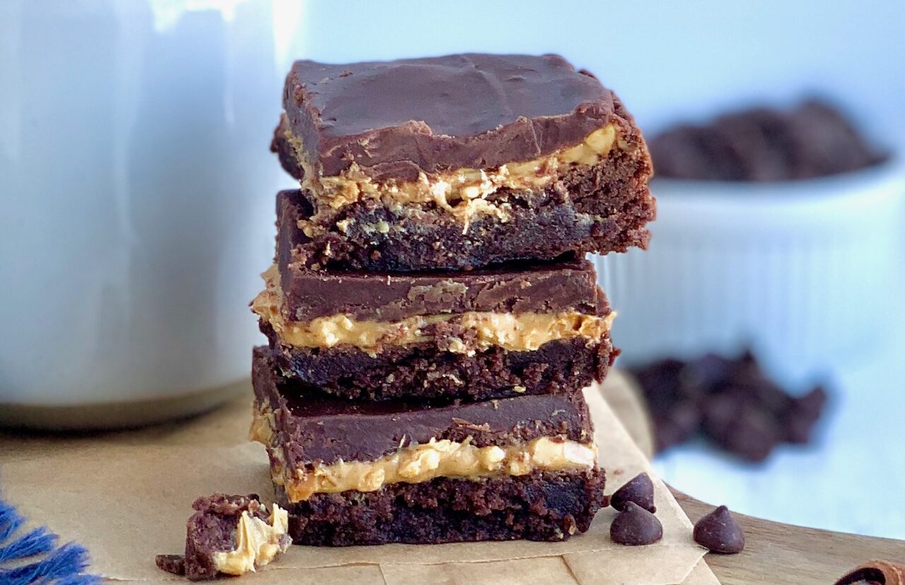 3 bars stacked up on each other next to a mug of coffee. Each bar ahs 3 layers-- a brownie layer on the bottom, then peanut butter, and topped with a chocolate marshmallow combination.