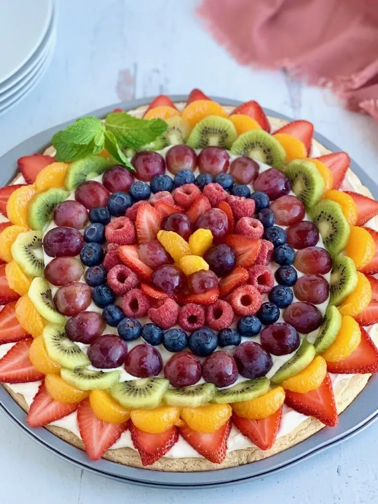 A large round sugar cookie crust with cream cheese and sliced fruit arranged in a colorful pattern. The fruit includes strawberries, mandarin oranges, kiwi, grapes, blueberries, and raspberries,