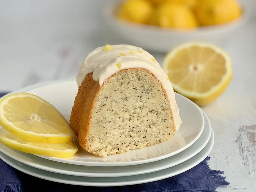 A slice of lemon poppy seed bunt cake with cream cheese frosting on a serving plate with 2 thin slices of lemon.