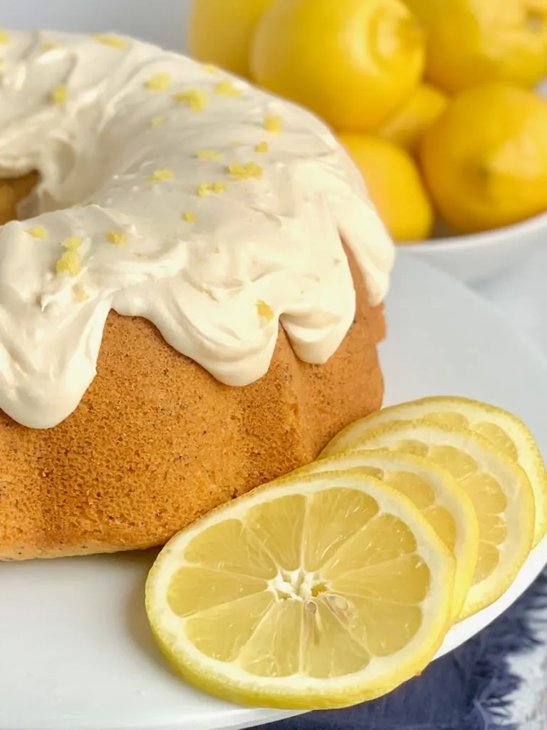 A close up of a lemon bundt cake with lemon cream cheese frosting smothered over the top of it on a cake plate with 4 thin slices of lemon on the side.