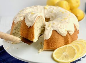 A lemon poppy seed bundt cake topped with a creamy lemon cream cheese frosting on a cake plate. A cake server is lifting a big slice off of the plate.